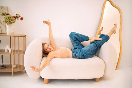 Female model is comfortably lying on a plush sofa, playfully stretching in a bright room with modern decor and a large mirror, embodying a carefree atmosphere, whiteの写真素材