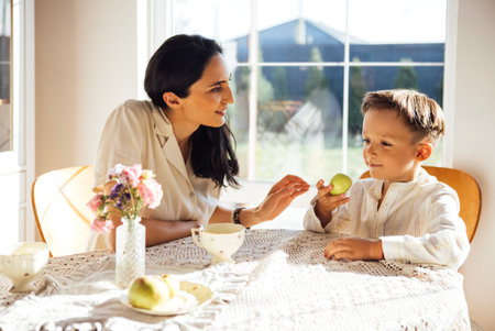 A woman and a young boy are sharing a green apple at a sunlit table, surrounded by flowers and tea cups, capturing a heartwarming family momentの写真素材