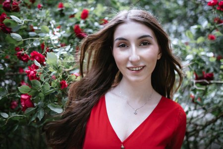 Female with long brown hair, dressed in red blouse, smiles in a garden surrounded by blooming red...