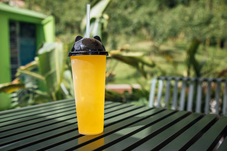 Yellow drink in a transparent cup with a black lid and straw on a green striped table, with vibrant foliage in the backgroundの写真素材