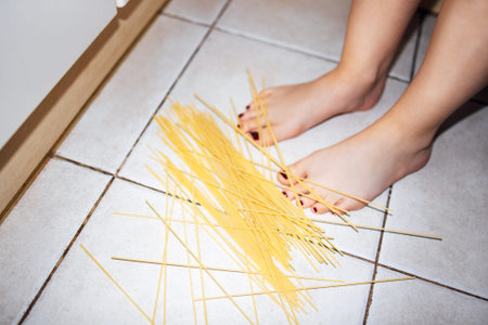 Person's bare feet are positioned on a tiled kitchen floor with spaghetti scattered around, highlighting a domestic environmentの写真素材