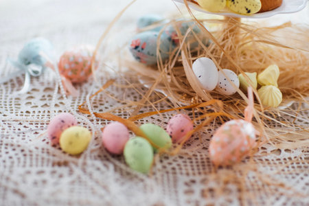 Decorative Easter eggs in various pastel colors are scattered on a lace tablecloth with straw,...