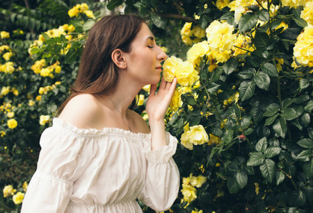 Female in white dress is interacting with vibrant yellow roses in a garden, surrounded by greenery, embodying tranquility and connection with natureの写真素材