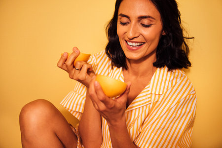 Happy woman with dark hair, dressed in striped pajamas, is holding fresh fruit, radiating joy and positivity in a bright yellow setting, showcasing a cheerful atmosphereの写真素材
