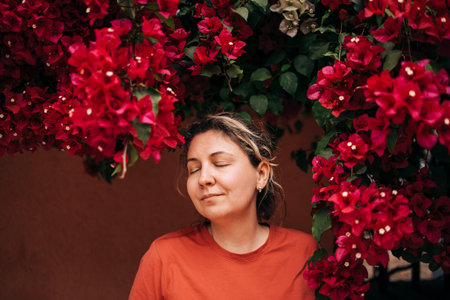 Female enjoying peaceful moment with closed eyes, framed by lush red bougainvillea flowers, creating a serene and calming atmosphere in natureの写真素材