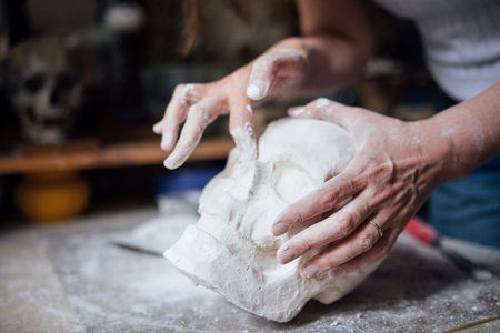 Artisan's hands are skillfully shaping a plaster skull model on a workbench, surrounded by tools and materials, highlighting the intricate art of sculpture creationの写真素材