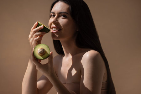 Female model with dark hair holds avocado halves, smiling and promoting healthy eating, with soft lighting and a neutral background enhancing the natural aestheticの写真素材