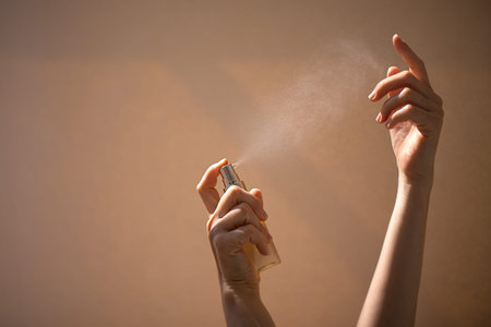 Hands of a woman spray mist from a bottle, creating a fine mist against a soft, neutral background,...