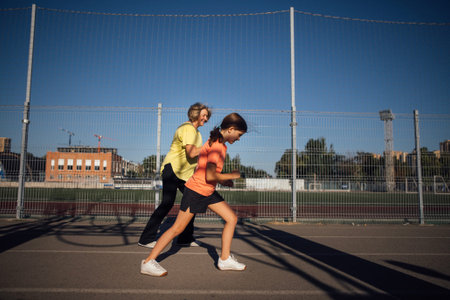 Senior woman supports young girl as they run together on an outdoor track, with a sports field and...