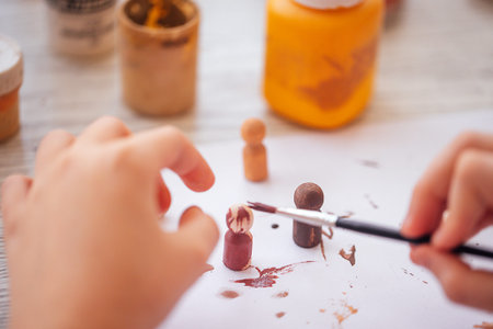 Child's hand applies paint to wooden figurines on a sheet of paper, with jars of paint visible on a...