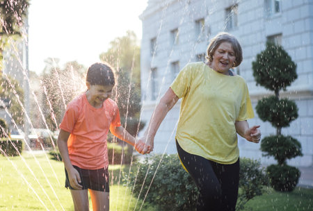 Elderly woman and little girl hold hands while playing in water spray, surrounded by greenery and a...