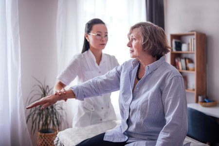 Asian woman physiotherapist helps senior female patient with arm exercises in a well-lit therapy room featuring indoor plants and a cozy atmosphereの写真素材