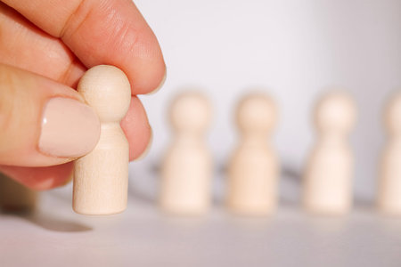 Hand holds a wooden figurine in front of a line of identical wooden figures on a neutral background