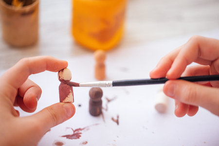 Hands of a child apply paint to wooden figurines using a brush, with jars of paint in the background...