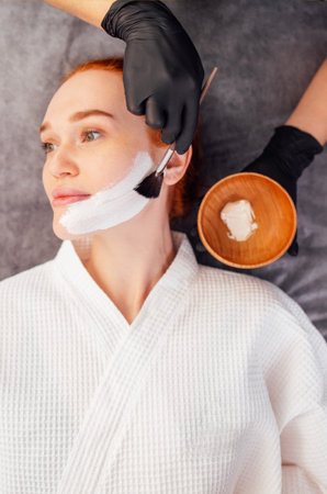 Woman enjoying a facial treatment with white cream on her face, lying on a soft surface,...
