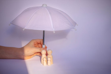 Hand supports a white umbrella above three wooden figurines, symbolizing family, set against a soft...