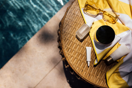 Skincare items placed on a wicker table next to a swimming pool, accompanied by sunglasses and a yellow striped towel, creating a summer vibeの写真素材