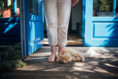 Woman with bare feet stands on a wooden porch, an orange cat lies comfortably at her feet, with blue...