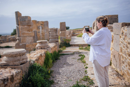 Woman wearing a white shirt photographs ancient stone ruins with columns at an archaeological site, Volubilis, Morocco,  surrounded by greenery and gravel pathwaysの写真素材