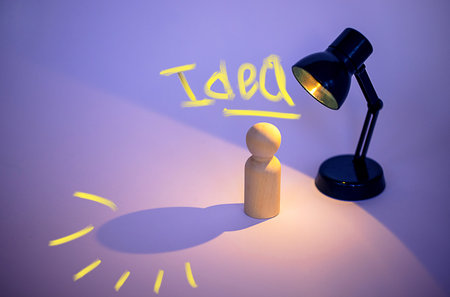 Wooden figurine stands under a desk lamp with the word "Idea" illuminated above, casting a shadow on a smooth surface in a workspaceの写真素材