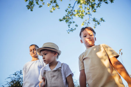 Three children, two boys and one girl, are standing together outdoors under a bright blue sky, surrounded by trees, capturing the essence of childhood joy and camaraderieの写真素材
