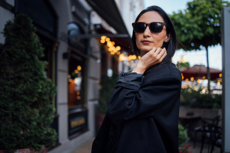 Confident woman in sunglasses poses outdoors, surrounded by lush greenery and soft ambient lights,...