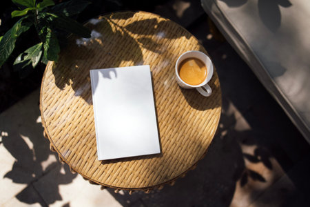 Blank notebook rests on a round wicker table next to a white coffee cup filled with coffee, with...