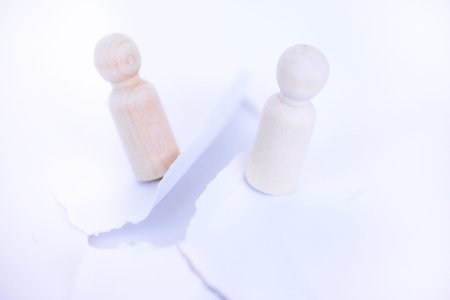 Two wooden figures stand apart, divided by a torn white paper piece, set against a soft light...