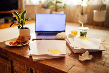 Workspace scene with a laptop, blank papers, and a plate of snacks on a wooden table, accompanied by...