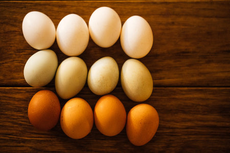 Assorted eggs in shades of white, brown, and green are arranged in a grid on a wooden surface,...