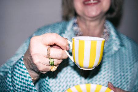 Mature woman presents a yellow striped teacup and bowl, adorned with jewelry, wearing a patterned...