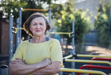 Older woman in yellow shirt poses with arms crossed in an outdoor fitness area, featuring exercise...