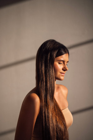 Female model with long dark hair stands in soft light, eyes closed, radiating tranquility and...