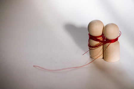 Two wooden figures are intertwined with red thread, standing closely on a light surface, with soft...