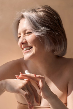 Woman with silver hair is joyfully applying skincare product on her hands, highlighting beauty routine in a serene, softly lit space with warm tonesの写真素材