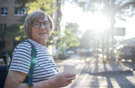 Senior woman with gray hair and glasses enjoys a coffee cup while walking along a sunlit street, surrounded by buildings and greenery