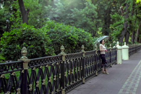 Beautiful young brunette standing with umbrella near fence in parkの写真素材