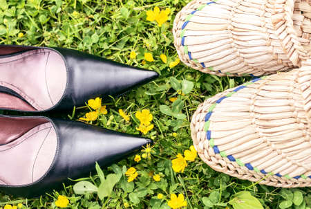 Black shoes and straw woven slippers on green grass like metaphore about town and countryの写真素材
