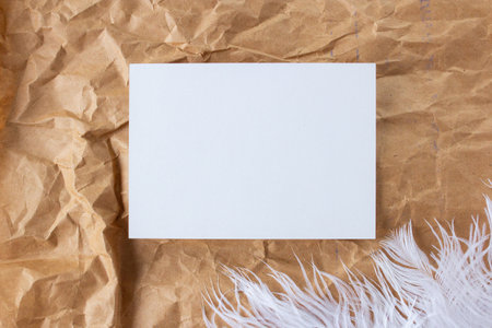 Blank greeting card on crumpled craft paper and ostrich feather flat lay, white empty invitation mock up with copy space on beige backgroundの写真素材
