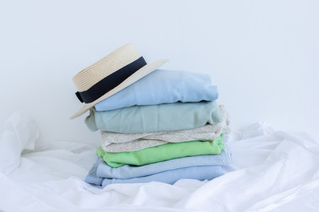 Pile of clothes with straw hat on white, pastel color palette of stacked sweaters and jeans, fresh clothing folded for ironing, fashion store concept with copy space, textile close upの写真素材