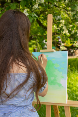 Woman artist with long hair painting plein air in spring park, wooden easel and canvas standing outdoor with green background, young girl in summer dress drawing, view from the backの写真素材