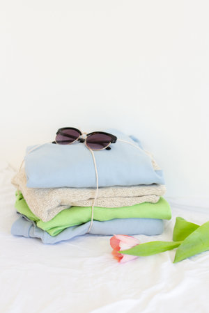 Pile of clothes, sunglasses and one pink tulip on white, pastel color palette of stacked sweaters...