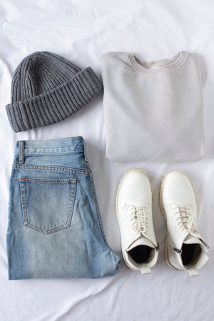 Female trendy clothes flat lay with jeans and sweatshirt on white sheet background, minimalistic travel set of clothes, women set of blue jeans, gray sweatshirt, white boots, gray knitted hat, fashion blogger content, autumn and spring look of the dayの写真素材