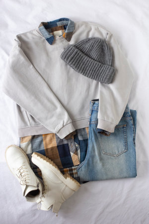 Travel set of clothes, Female trendy clothes flat lay on white sheet background, women set of blue jeans, gray sweatshirt, white boots, gray knitted hat and checked shirt view, fashion blogger content, autumn and spring look of the dayの写真素材