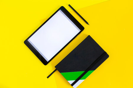 Home office flat lay with tablet and black notebook on yellow background