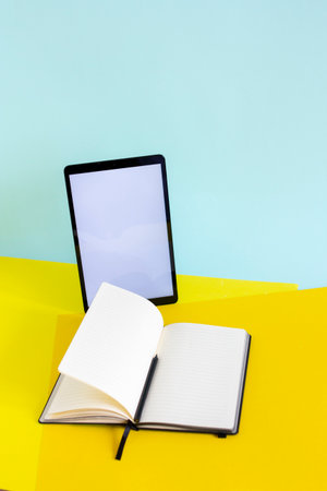 Blank tablet and ope notebook with pencil on yellow and blue background, work desk with copy space,...