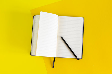 Open blank notebook and black pencil on yellow and blue background, white pages close up, school and...