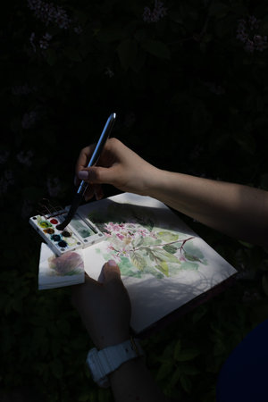 Watercolor painting of floral bird cherry tree in women hands. Female artist drawing in the dark of botanical garden and park, plein air. Summer sketching in shadowsの写真素材