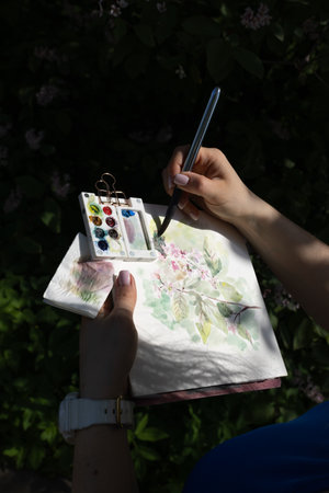 Watercolor painting of floral bird cherry tree in women hands. Female artist drawing in the dark of botanical garden and park, plein air. Summer sketching in shadowsの写真素材
