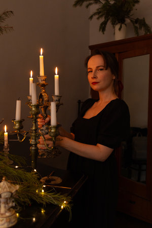 Beautiful young woman standing in candles in candlesticks in dark room. Portrait of girl in black dress in darkness, vertical shotの写真素材
